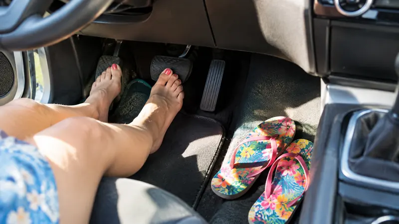 Nackte Füße neben den Pedalen eines Autos, daneben liegen Flip-Flops